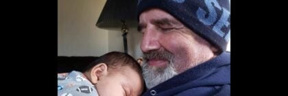 man with beanie, and dark blue sweatshirt sitting with an infant curled on his chest, a contented, loving smile on the mans face as he looks down at the baby.
