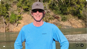 Sean O'Kane, smiling mane in hat, glasses, bright blue shirt in outdoor setting. 