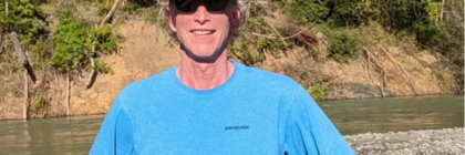 Sean O'Kane, smiling mane in hat, glasses, bright blue shirt in outdoor setting.